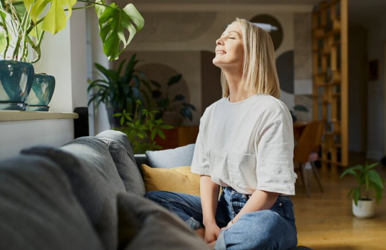 Woman sitting on a gray couch with eyes closed, basking in sunlight in a bright living room with plants nearby.